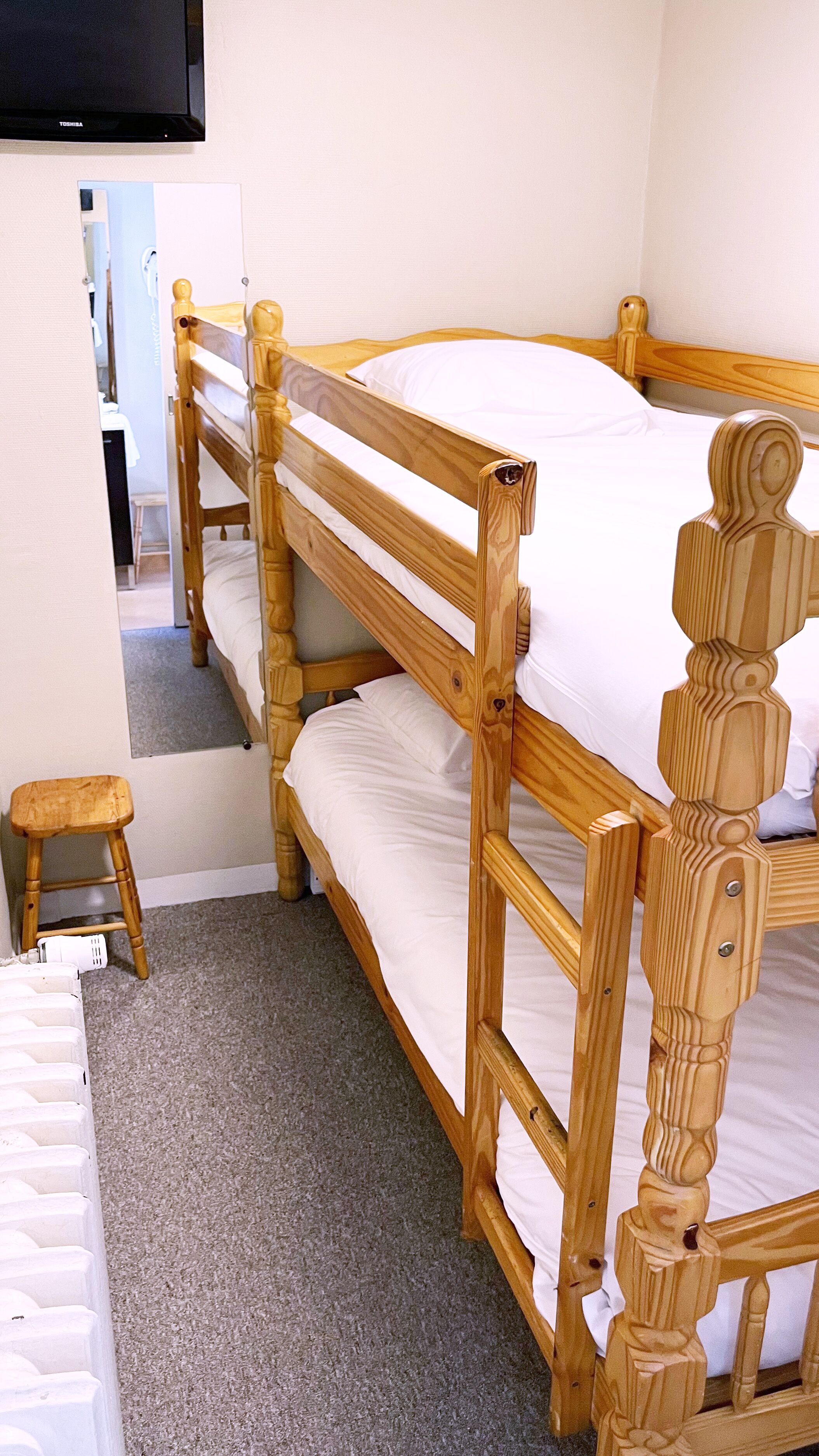 A bunk bed with a wooden frame and white bedding, a small wooden stool, and a television mounted on the wall.