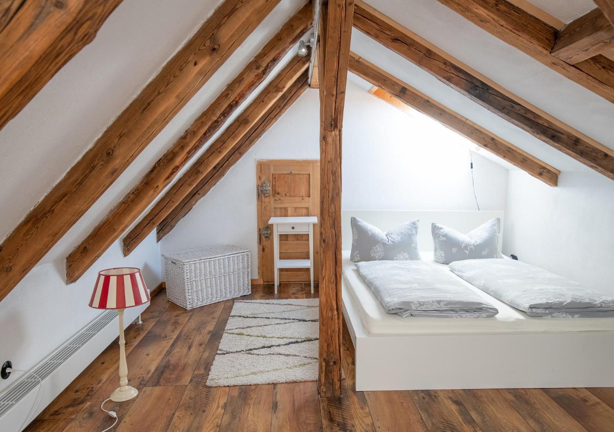 A bedroom with a sloped ceiling, wooden beams, a bed with pillows, a small table, a lamp, and a basket.