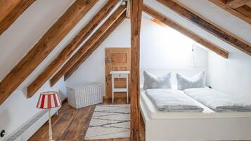 A bedroom with a sloped ceiling, wooden beams, a bed with pillows, a small table, a lamp, and a basket.