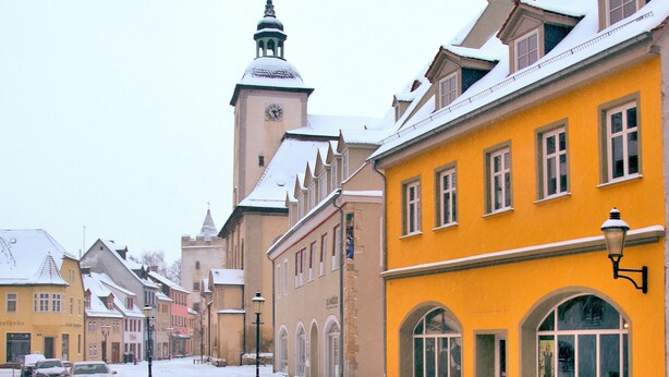 Una calle nevada con edificios históricos, incluyendo una torre destacada con un reloj.