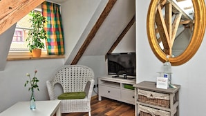 Un acogedor cuarto ático con techo de madera, una silla de mimbre blanca, un televisor, un espejo y una mesita con un jarrón de flores.