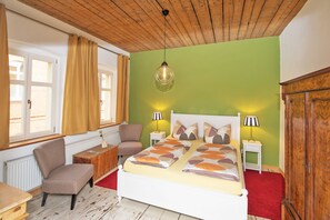 Un dormitorio con techo de madera, una cama con ropa de cama estampada, dos lámparas de noche, una silla, una mesita y un radiador.