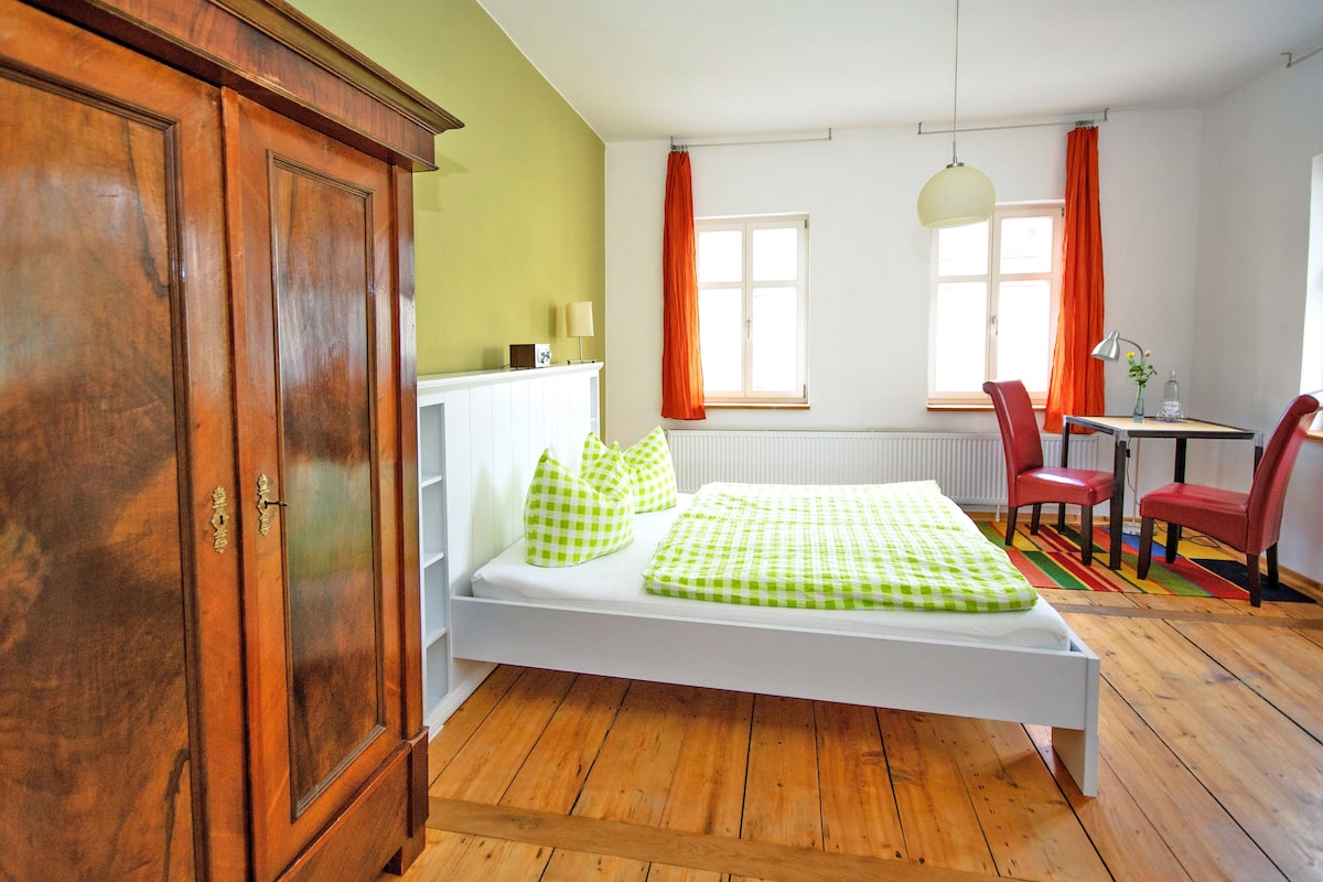 A room with a wooden floor, a white bed with green checkered bedding, a wooden wardrobe, a small table with a lamp, and a window with orange curtains.