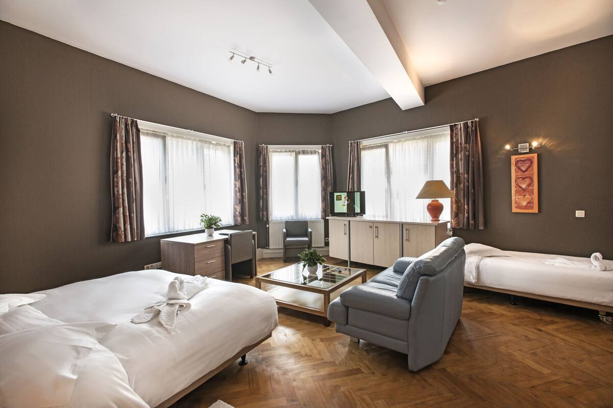 Habitación de hotel con cama, sofá, escritorio, silla, televisión y ventana con cortinas.