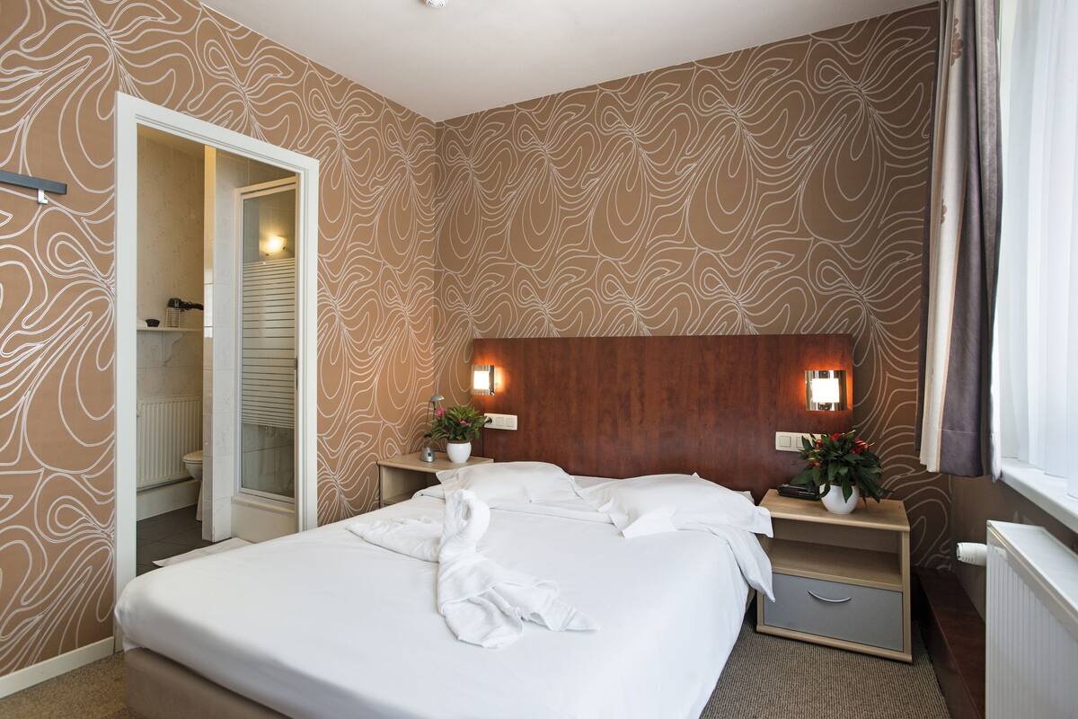 Habitación de hotel con una cama grande, dos mesitas de noche con lámparas, un cabecero de madera, un armario y un baño visible a través de una puerta abierta.