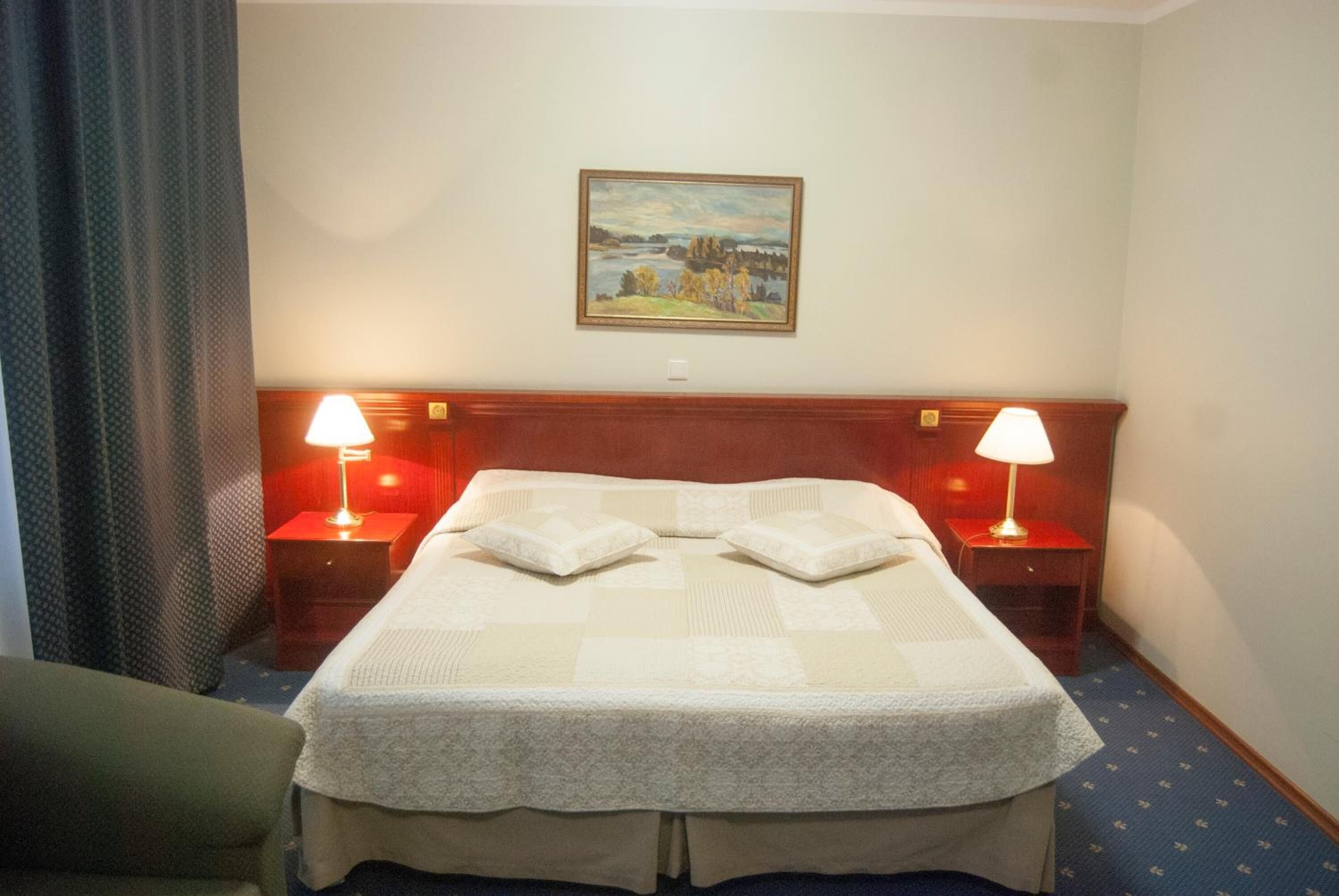A hotel room with a large bed, two bedside lamps, a painting on the wall, and a window with curtains.