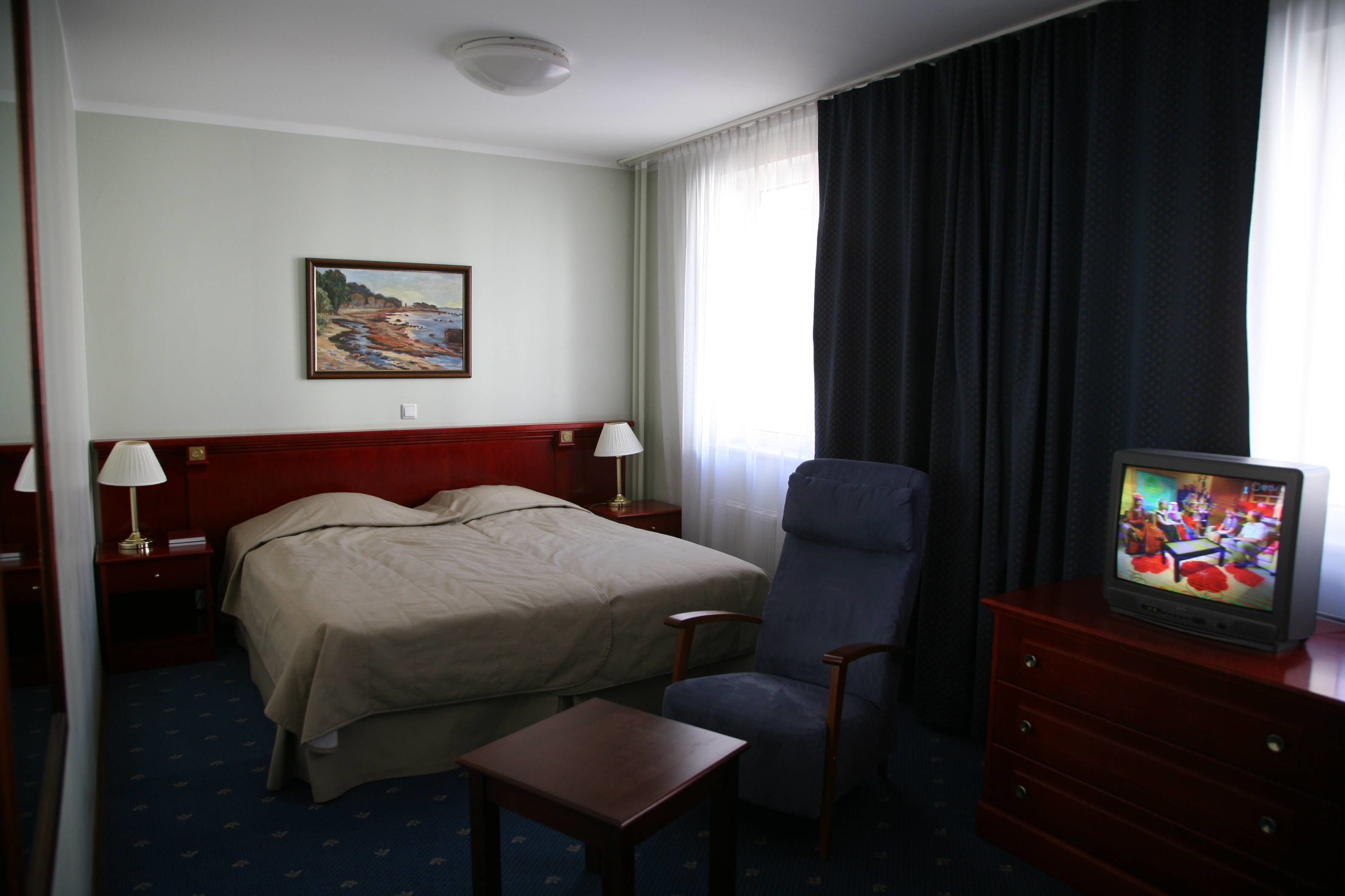 A hotel room with a bed, a chair, a TV, a desk, and a painting on the wall.