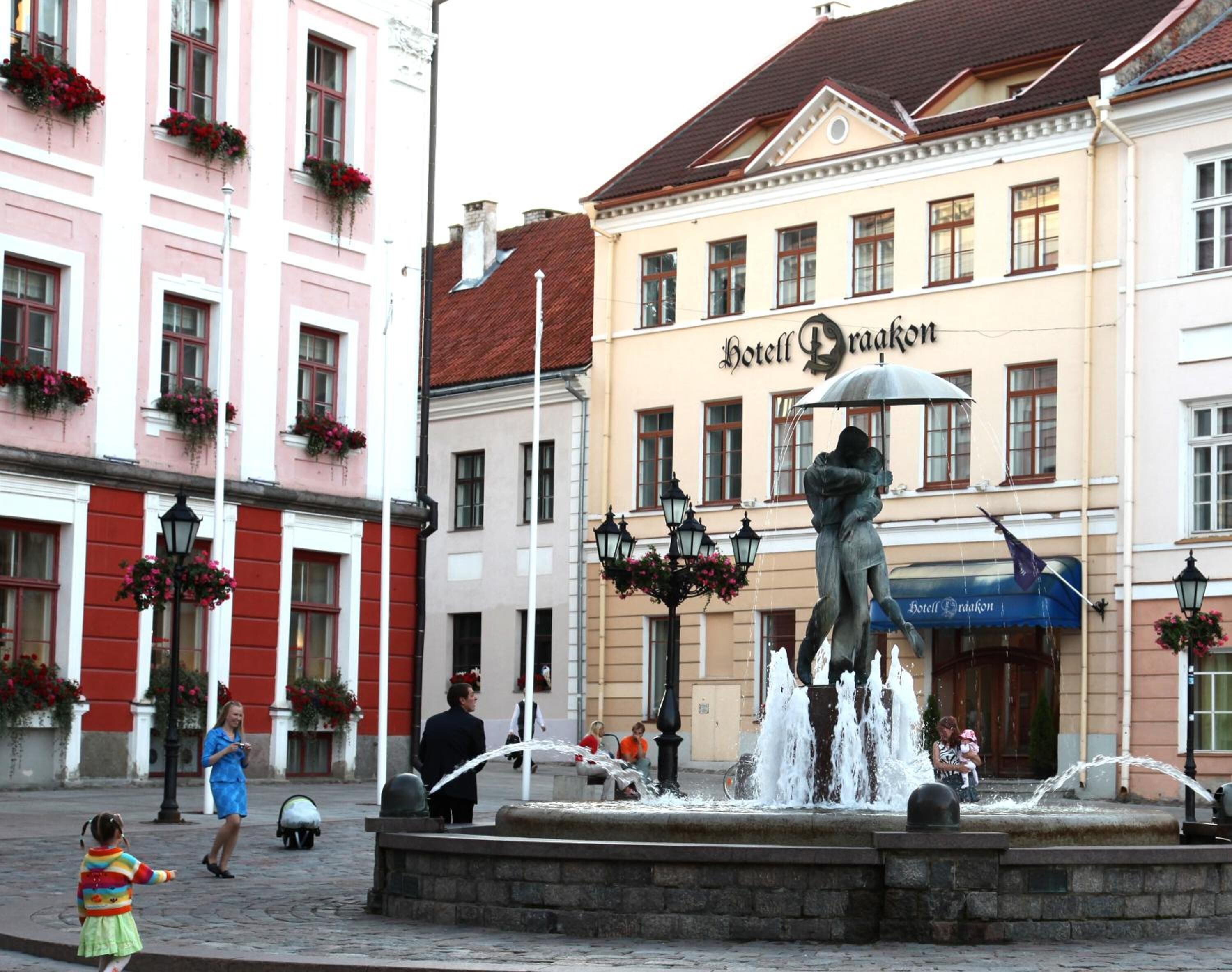 A public fountain with a statue in the center, surrounded by a stone basin. In the background, there is a building with the sign "Hotel Braakon".