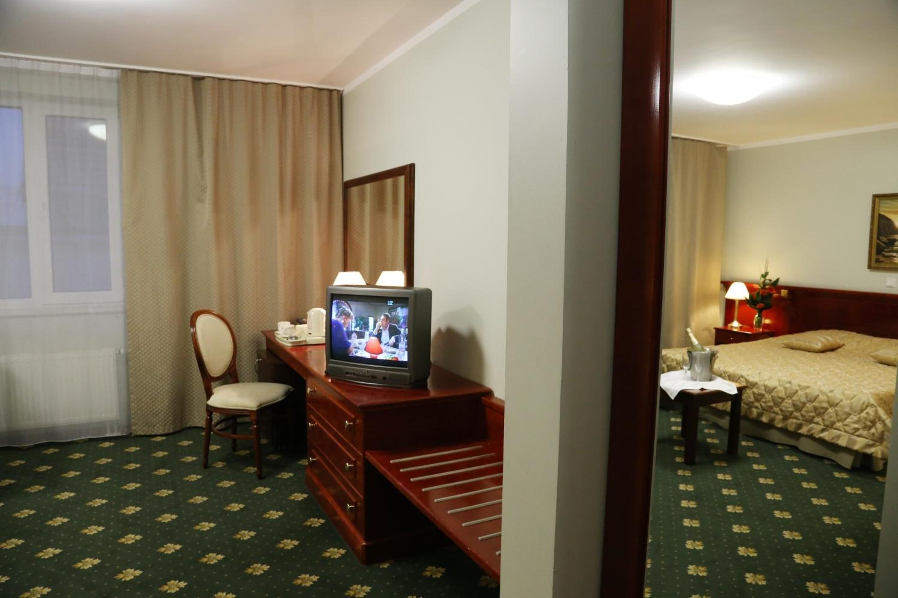Hotellihuoneessa on televisio, joka on sijoitettu puiseen lipastoon, beige värinen peitto on sängyllä, pieni pöytä, jossa on lamppu, ja tuoli.