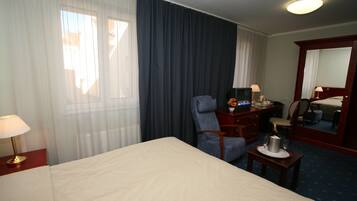 A hotel room with a large bed, a desk with a lamp, a chair, a small table with a bottle and glasses, and a mirror.
