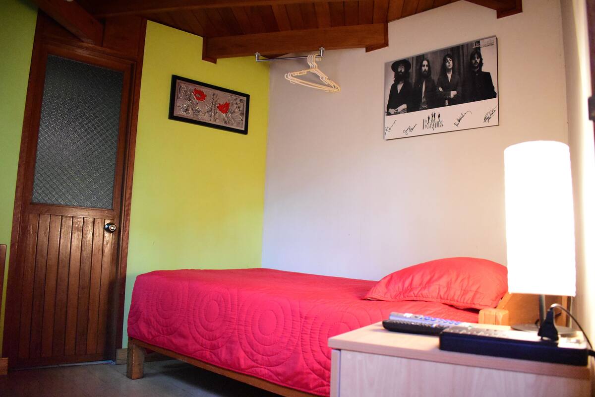 A bedroom with a bed, a nightstand, a lamp, a door, and a wall with a framed picture and a poster.