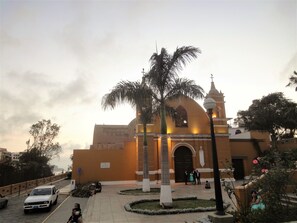 A church with a dome and a bell tower, surrounded by a paved area with palm trees and a few people.