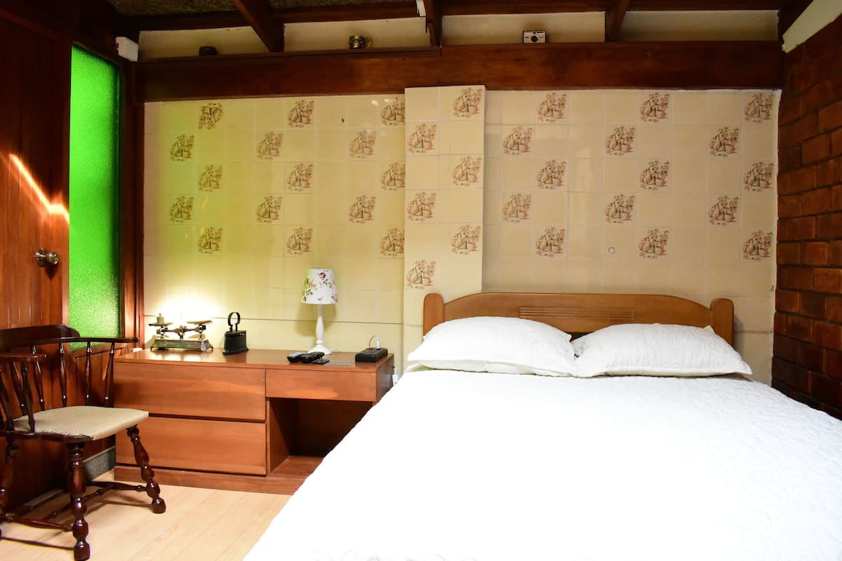 A bedroom with a wooden bed, a desk with a chair, and a green-framed window.