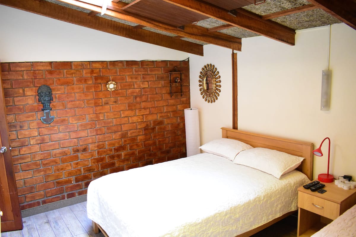 A bedroom with a brick wall, a wooden bed frame, a bedside table with a lamp, and a wall clock.