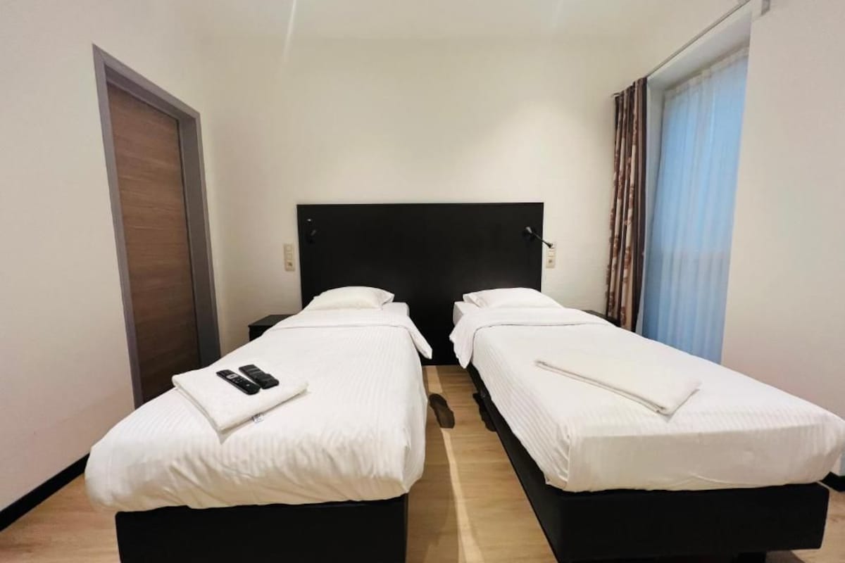 A hotel room with two single beds, a wooden floor, a black headboard, and a window with curtains.