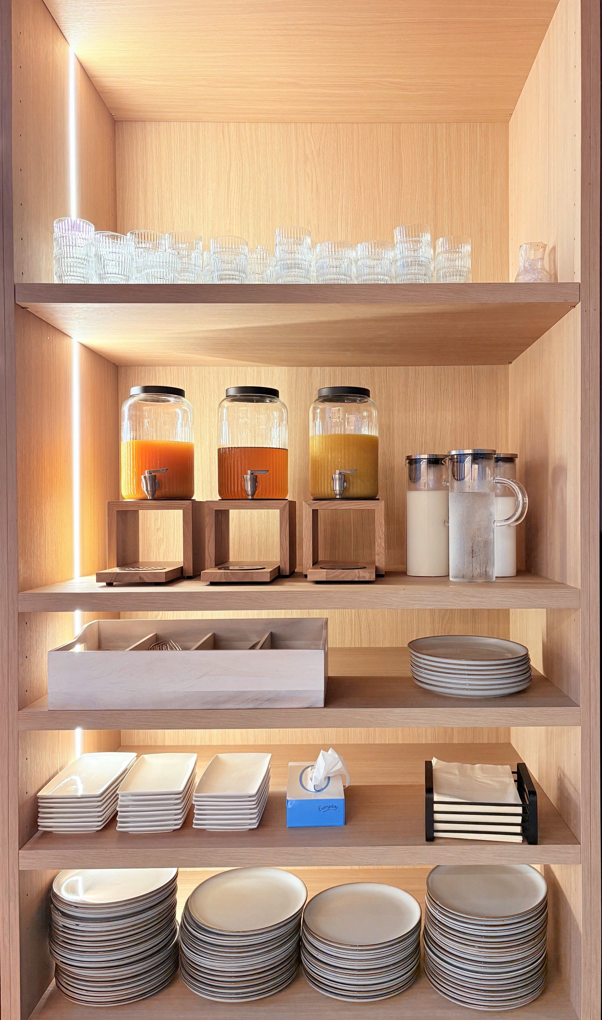 A wooden shelf unit with glass carafes, a stack of plates, and a tissue box.