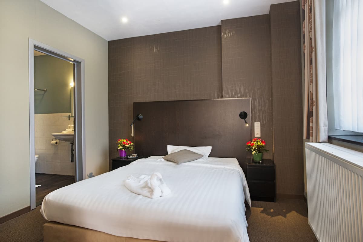 Ein Hotelzimmer mit einem großen Bett, einem Nachttisch mit Lampe, einem Fenster mit Vorhängen und einem durch eine offene Tür einsehbaren Badezimmer.
