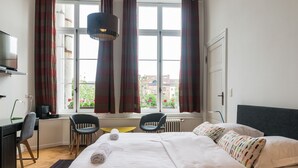 Una habitación de hotel con una cama, dos sillas, un escritorio, una lámpara y un ventanal con cortinas.
