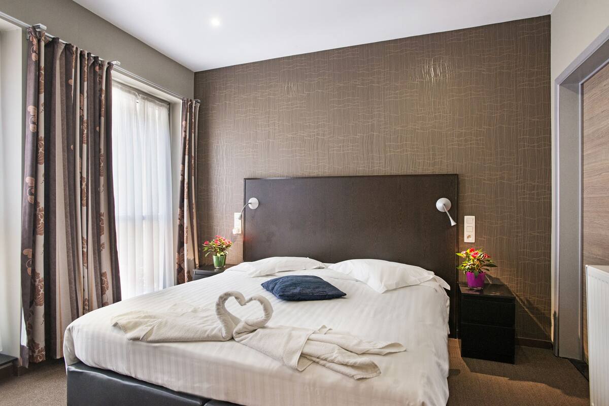 Ein Hotelzimmer mit einem großen Bett, einem Kopfteil, zwei Nachttischen mit Lampen, einem Fenster mit Vorhängen und einer Wand mit einer strukturierten Oberfläche.
