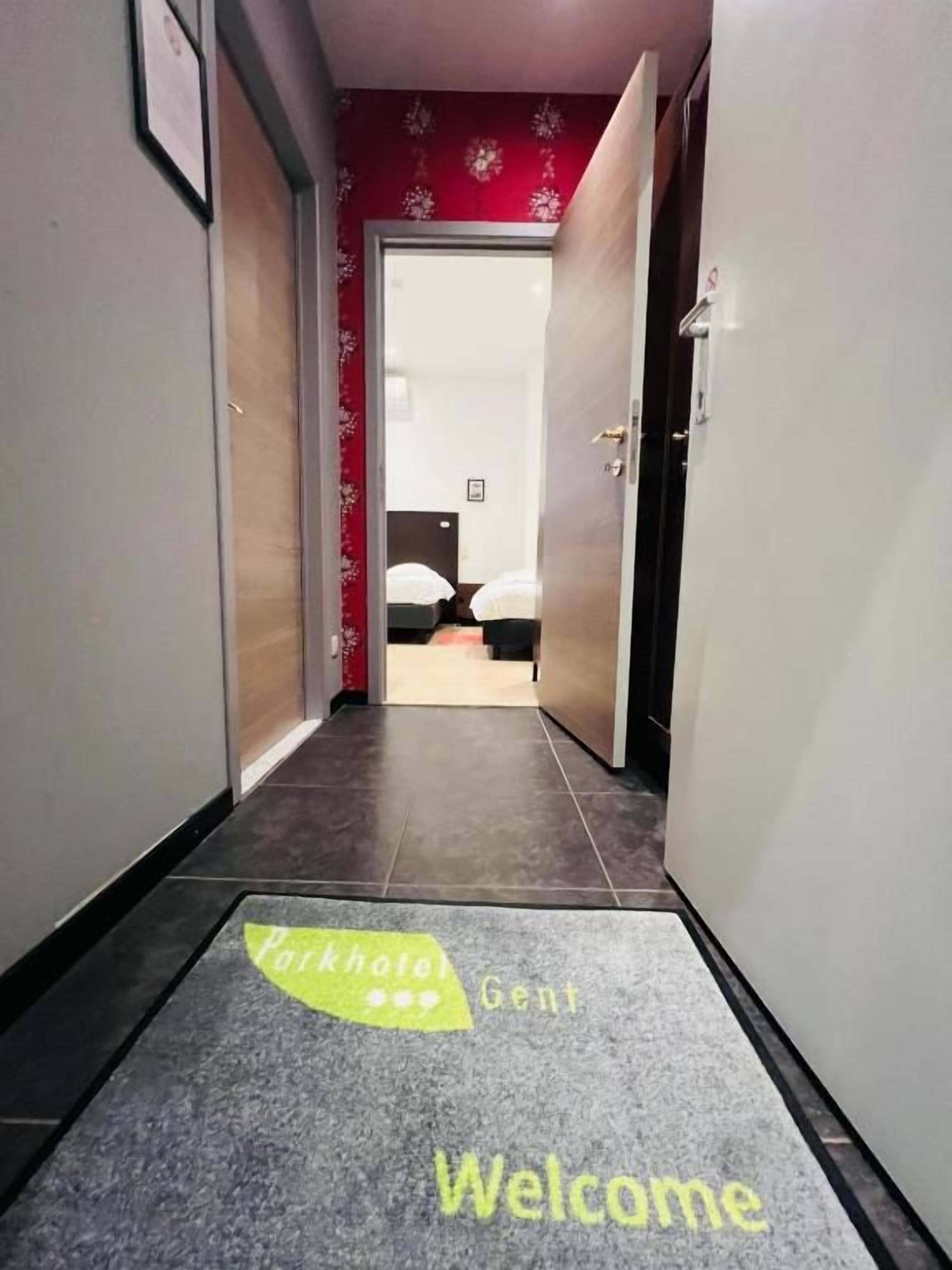 Un couloir d’hôtel avec un paillasson, une porte donnant sur une chambre et un mur rouge orné d’un papier peint à motifs floraux.