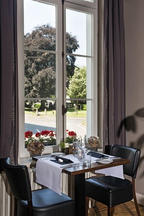 A dining area with a table set for two, a window with curtains, and a view of a park. - Parkhotel Gent Centrum (Ghent)