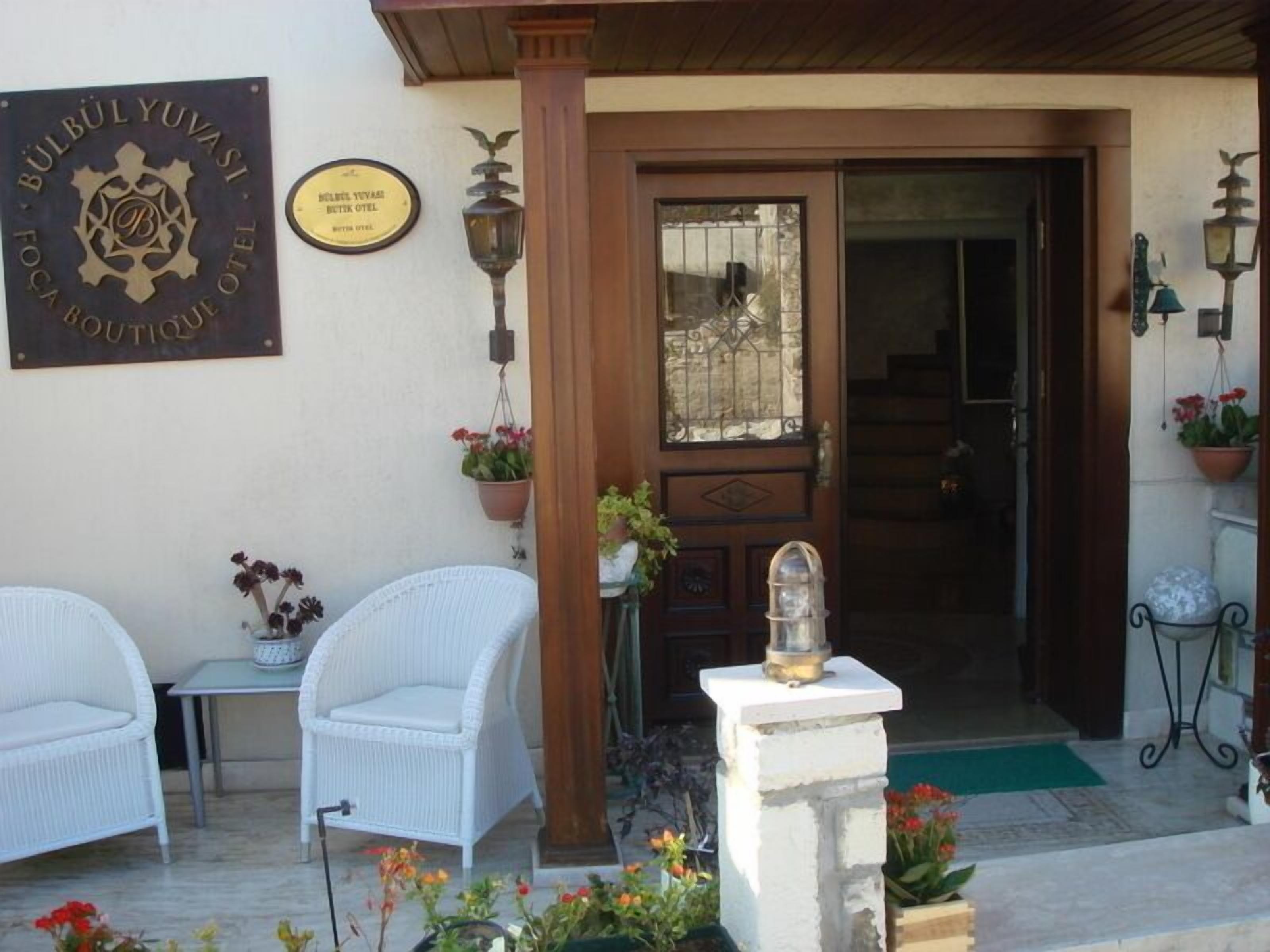 A hotel entrance with a wooden door, a signboard, and two white wicker chairs outside.