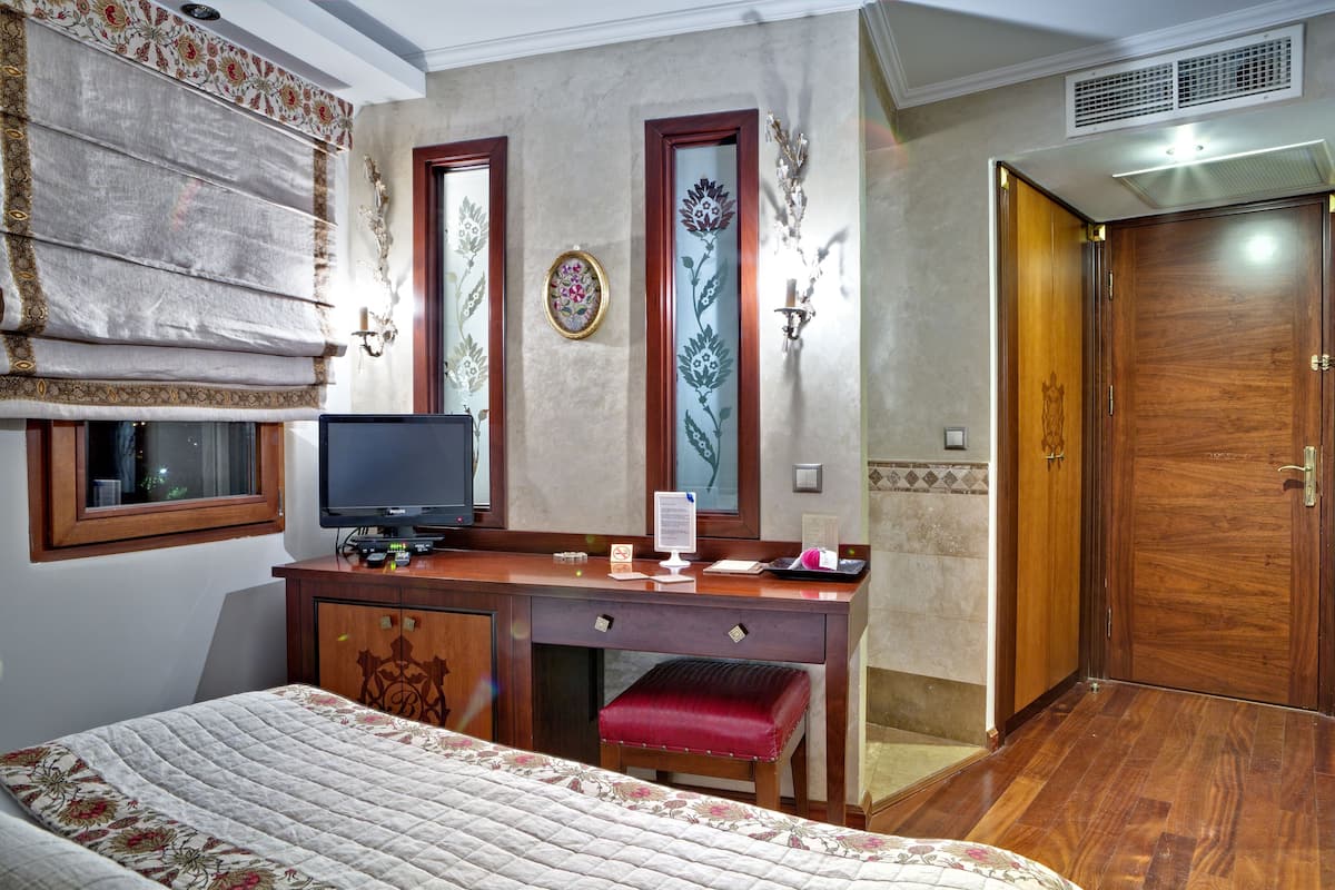A hotel room with a bed, a desk with a computer, a red bench, a wooden wardrobe, and a door.