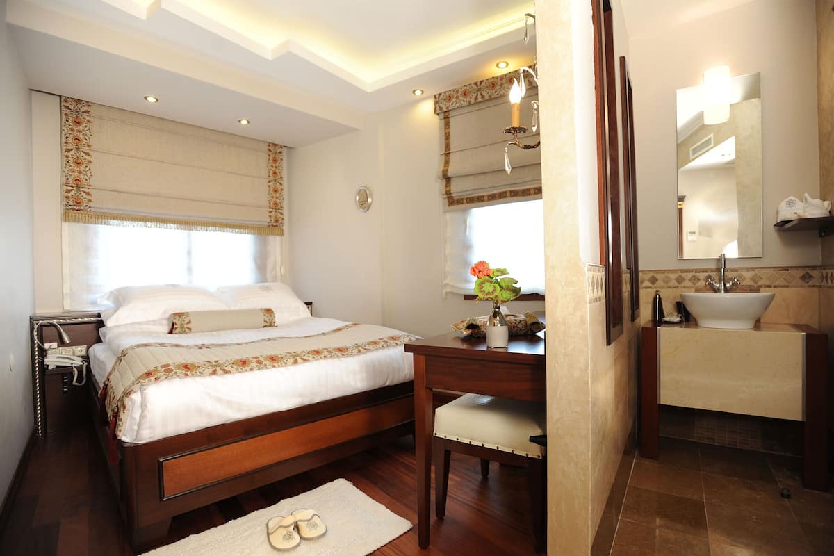 A bedroom with a wooden bed, bedside table, and a desk with a chair. There is a bathroom with a sink and mirror.