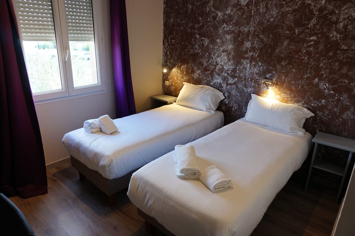 Ein Hotelzimmer mit zwei Betten, jeweils mit weißer Bettwäsche und Handtüchern, einem Nachttisch mit Lampe und einem Fenster mit Jalousien.