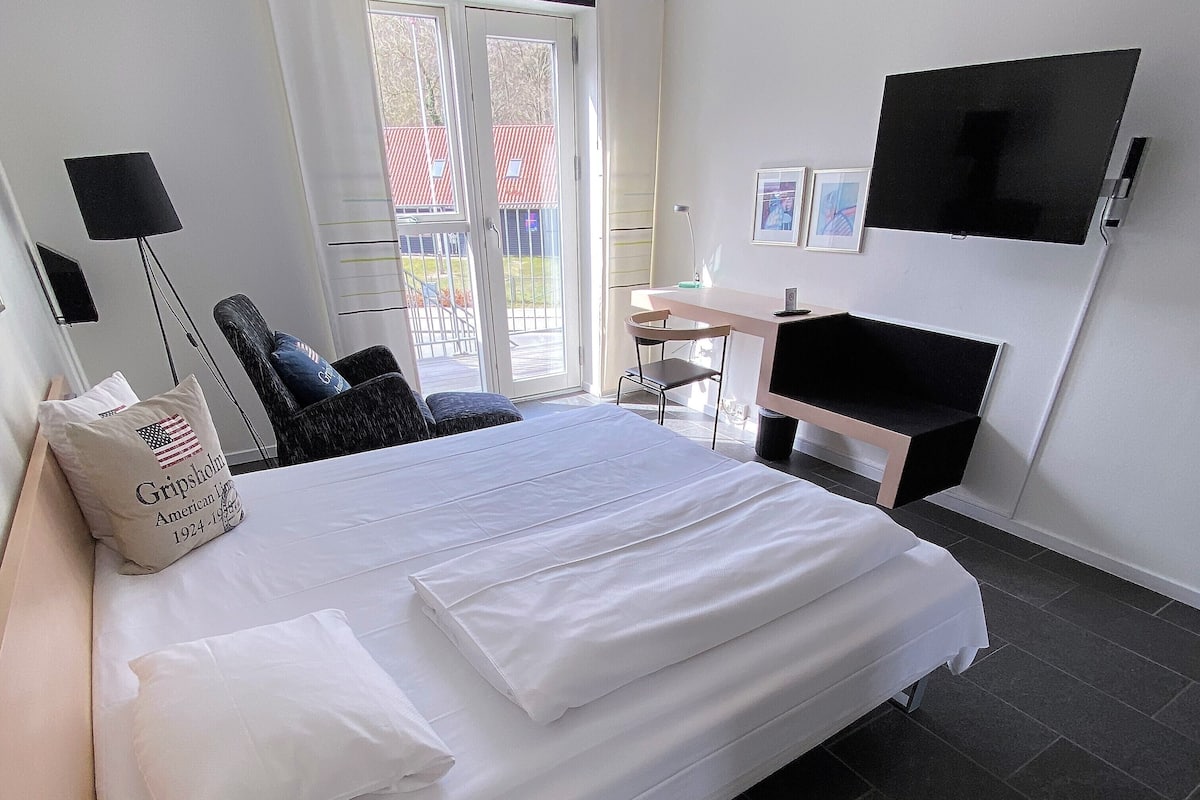 A hotel room with a large bed, a desk, a chair, a TV mounted on the wall, and a window with a view of the outside.