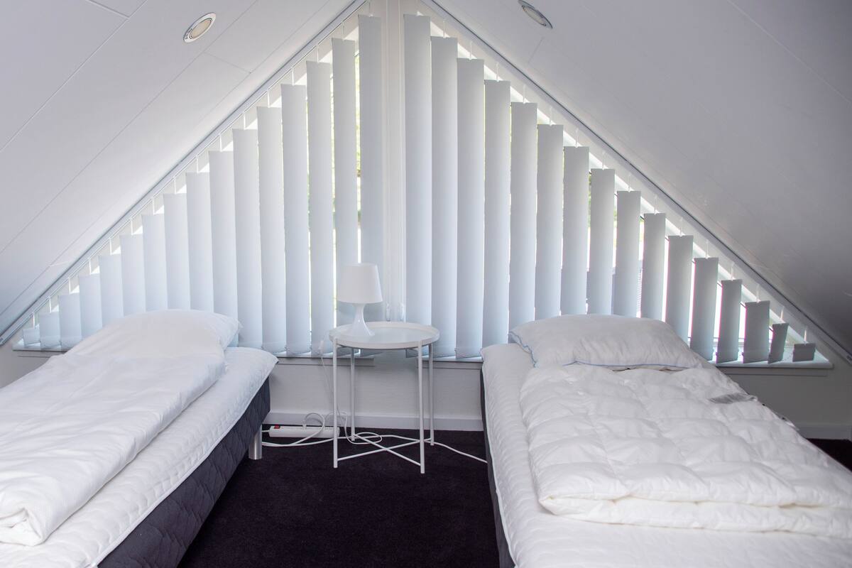 A small, triangular attic bedroom with two single beds, a small table, and a window with vertical blinds.