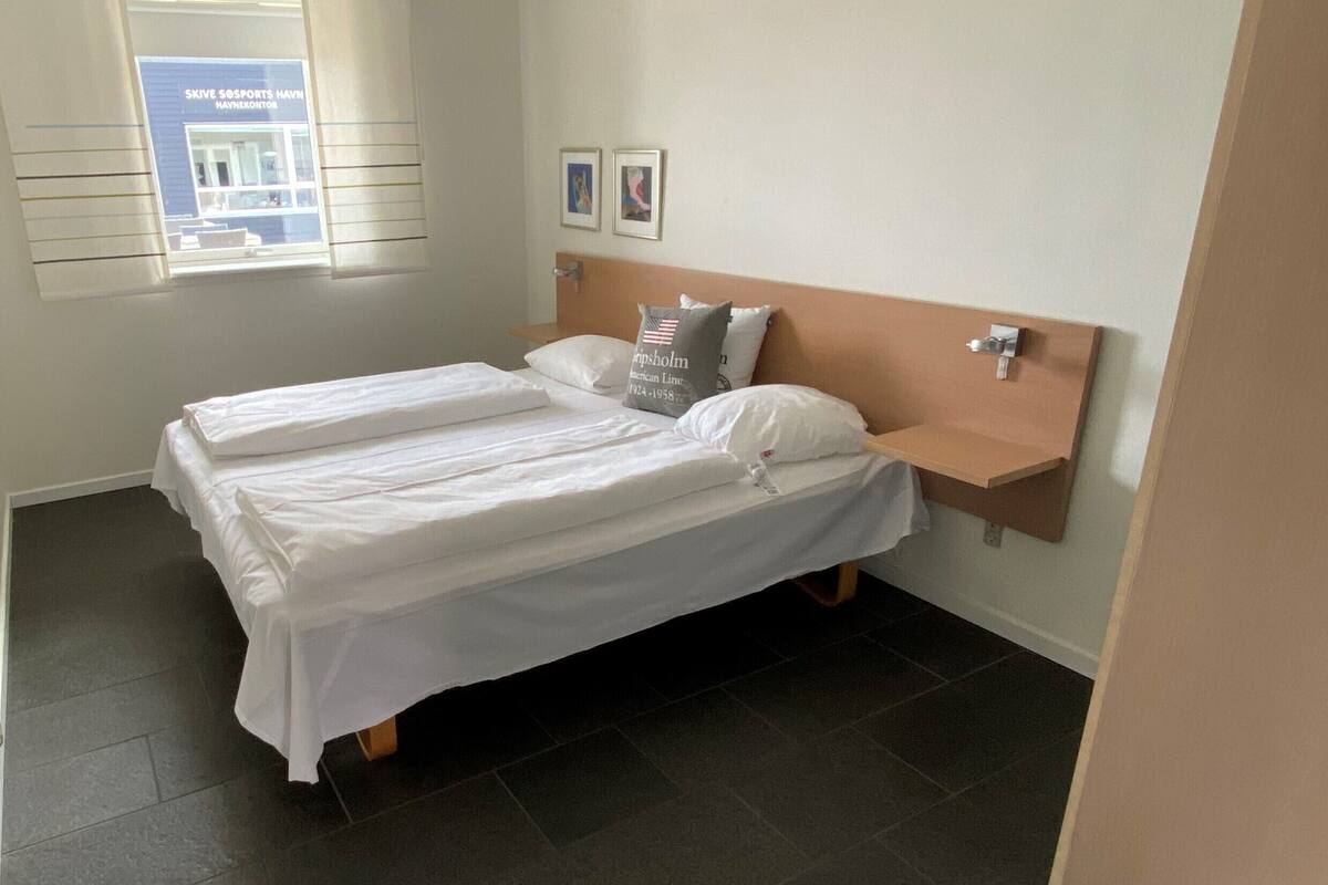A double bed with white linens, a wooden headboard, and a small shelf with a pillow.