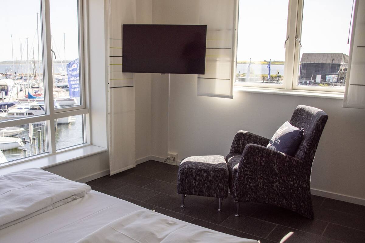 A hotel room with a large window offering a marina view, a flat-screen TV mounted on the wall, and a dark-patterned armchair.