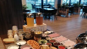 A buffet setup with various food items, including fruits, meats, and beverages, in a room with large windows and a view of the city. - Hotel Strandtangen (Skive)