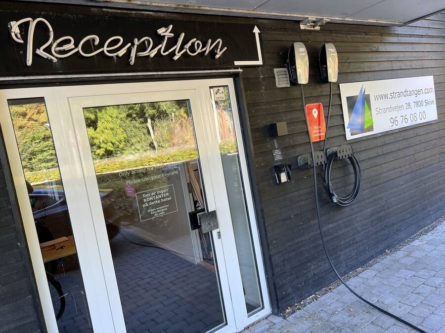 Un espace d’accueil avec un panneau indiquant qu’il est réservé aux clients de l’hôtel, une borne de recharge pour véhicules Tesla et un numéro de téléphone à contacter pour joindre l’hôtel.