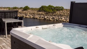 A hot tub with a wooden deck and a table with chairs, overlooking a rocky shoreline with greenery. - Hotel Strandtangen (Skive)