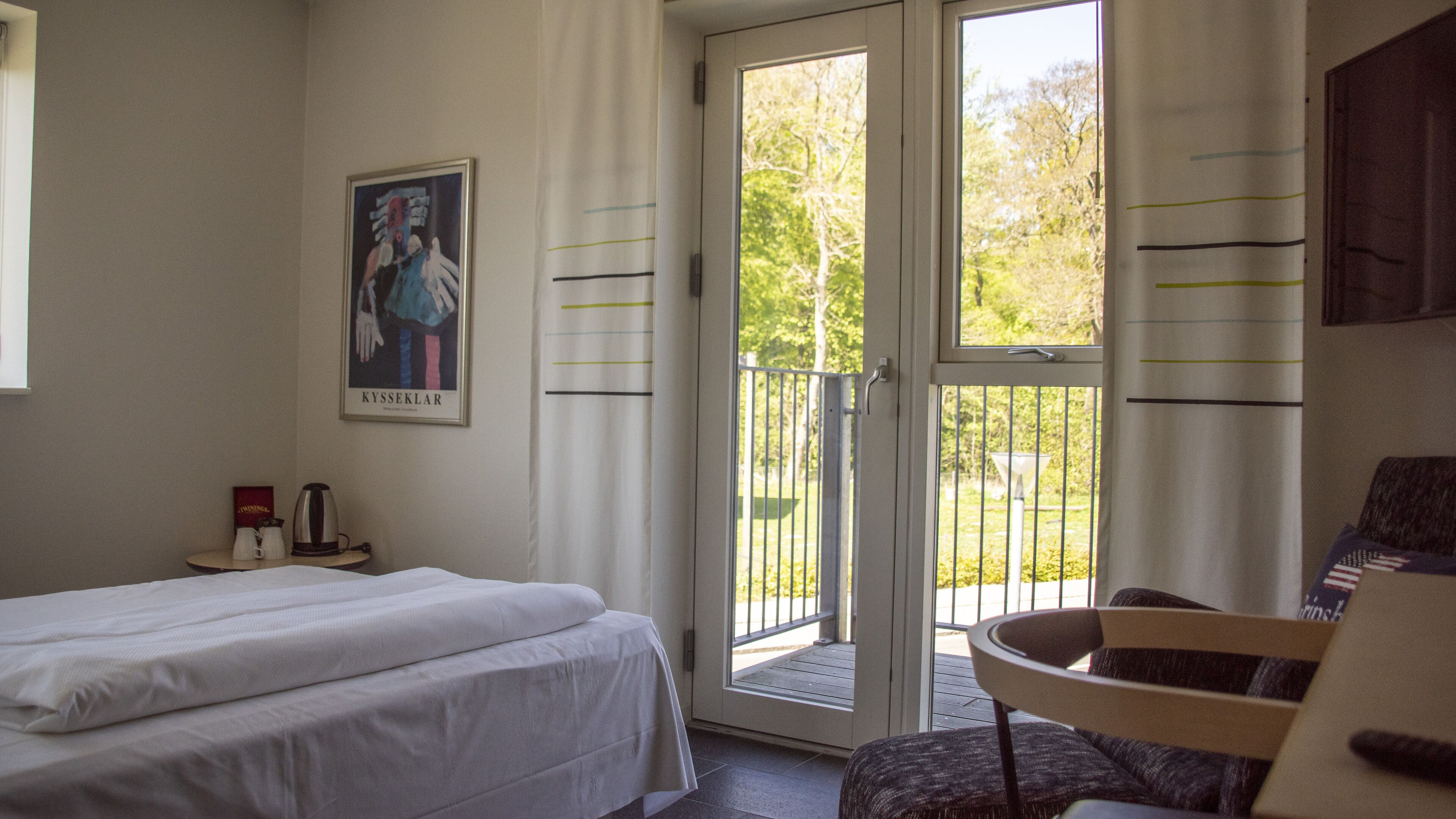 A hotel room with a bed, a chair, a window with a view of trees, and a poster on the wall.