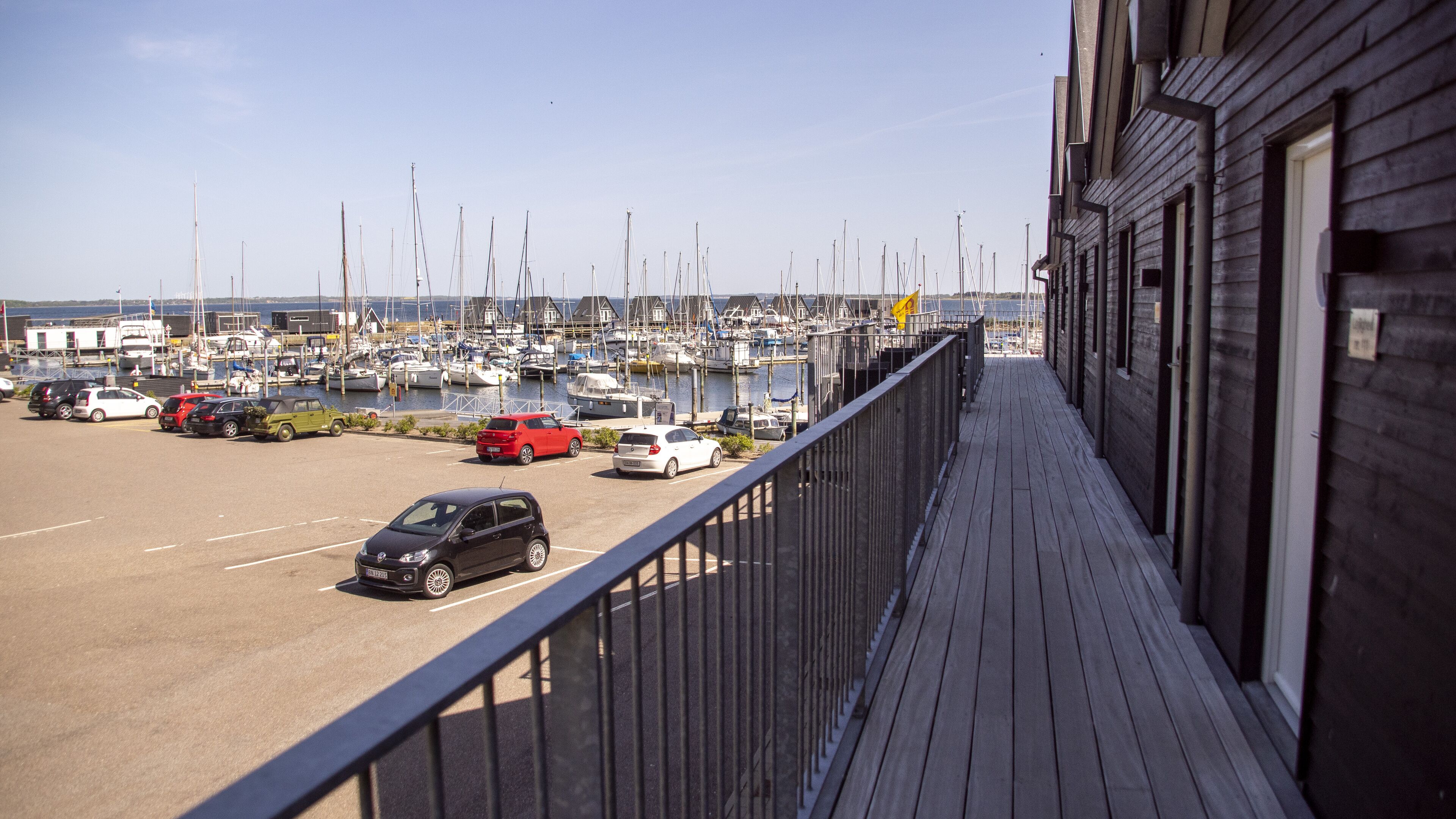 Un puerto deportivo con barcos amarrados, un estacionamiento con coches y un edificio de madera con balcón que da al puerto.