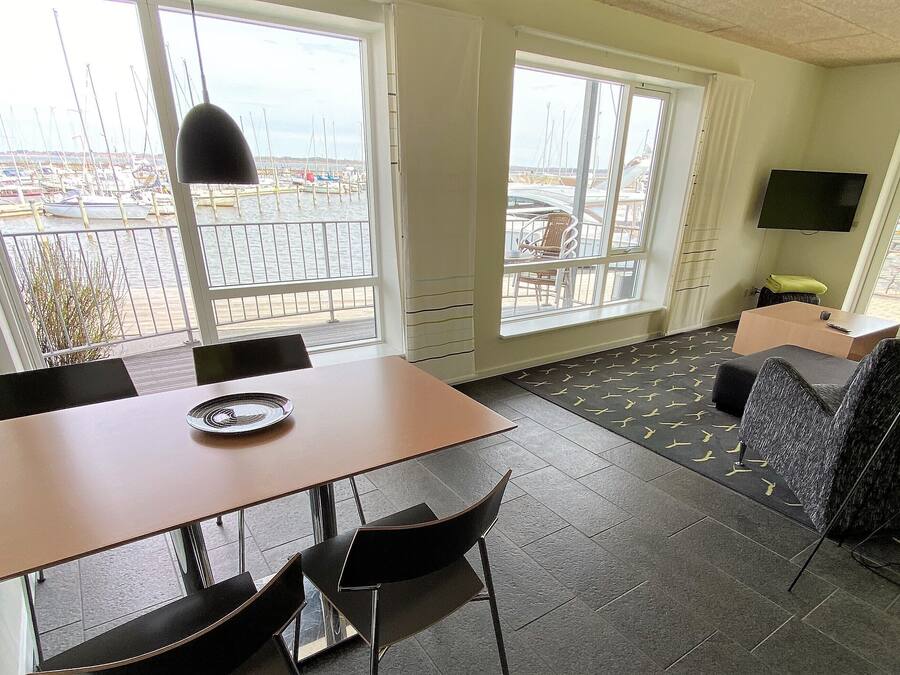 Una camera moderna con un tavolo da pranzo, sedie, un divano e una TV. Ampie finestre offrono vista su barche e un porto turistico.