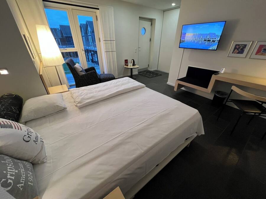 A hotel room with a bed, a desk, a chair, a TV, and a view of buildings through the window.