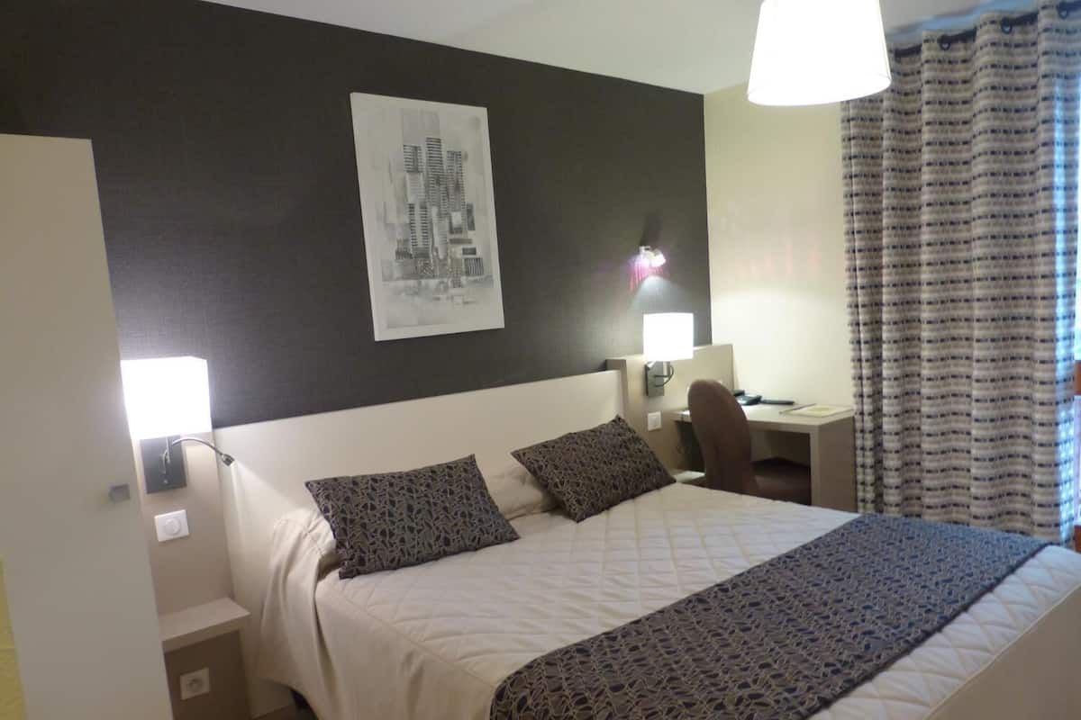 A hotel room with a bed, two pillows, a desk, a chair, a lamp, a wall-mounted light, and a window with curtains.