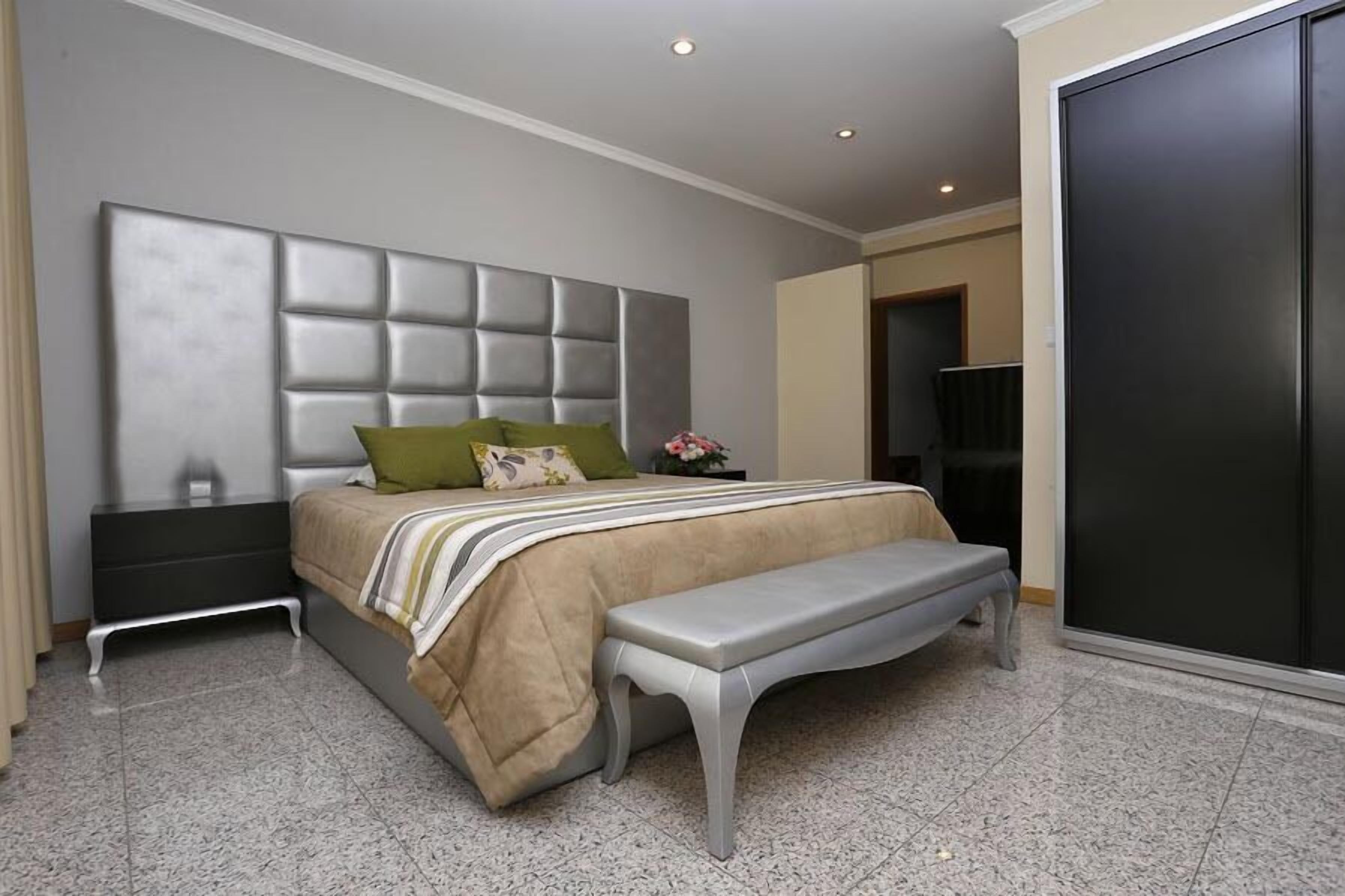 Un dormitorio moderno con un cabecero capitonado, una cama con edredón a rayas, un banco y un armario oscuro.