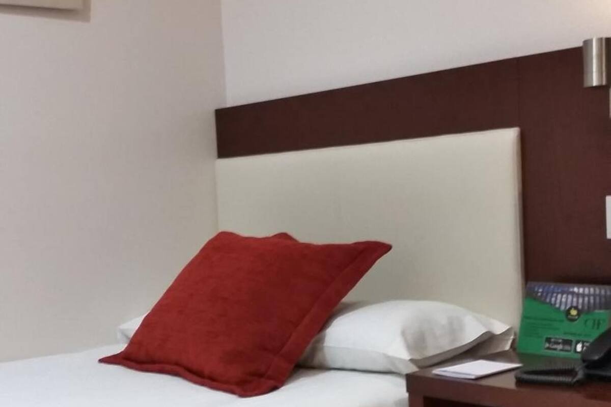 Una cama bien hecha con ropa blanca y una almohada roja, una mesita de noche de madera con una caja verde y un objeto negro, y una lámpara fijada a la pared.