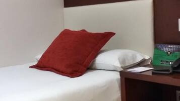 Quarto de hotel com uma cama, um travesseiro vermelho, um travesseiro branco, uma mesinha de cabeceira com abajur e uma caixa verde sobre a mesinha.