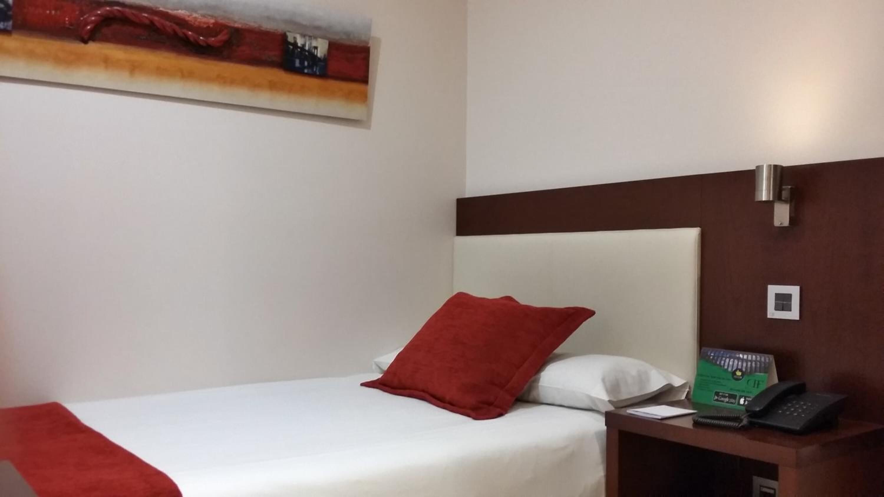 A hotel room with a bed, a red pillow, a white pillow, a nightstand with a telephone, and a painting on the wall.