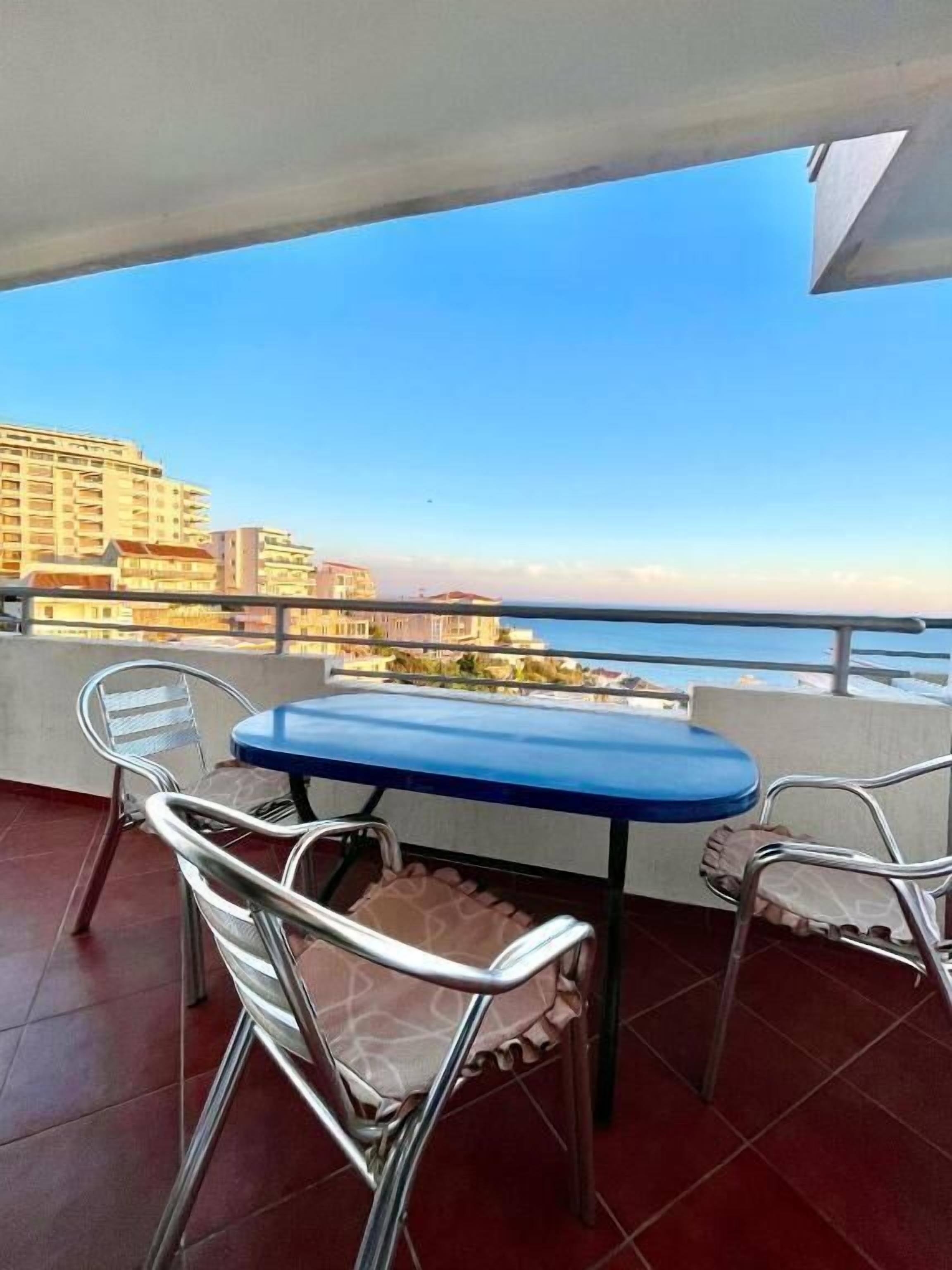En balkon med et bord og stole, med udsigt over et kystområde med bygninger i baggrunden.