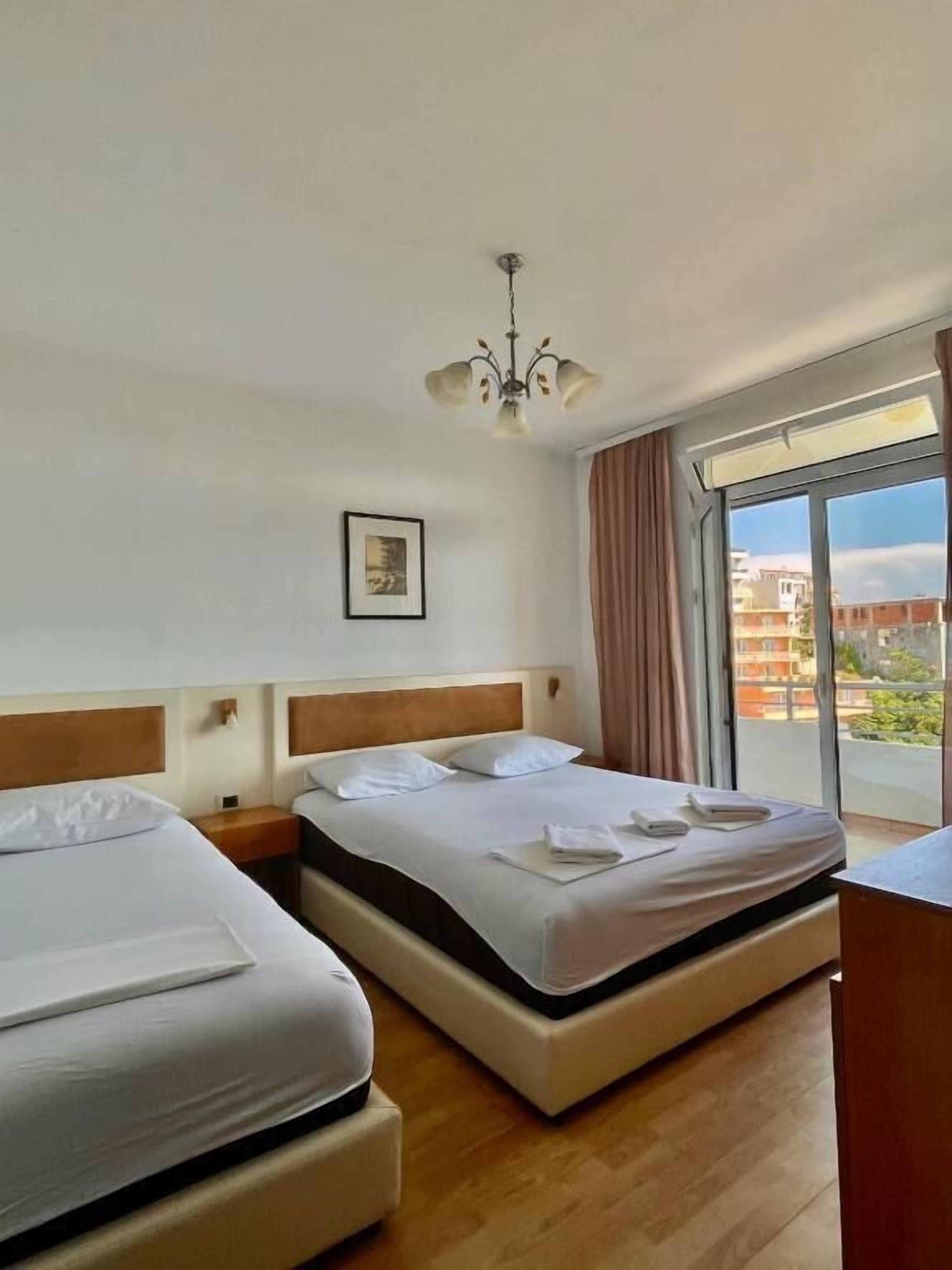 A hotel room with two beds, a chandelier, a large window with curtains, and a framed picture on the wall.