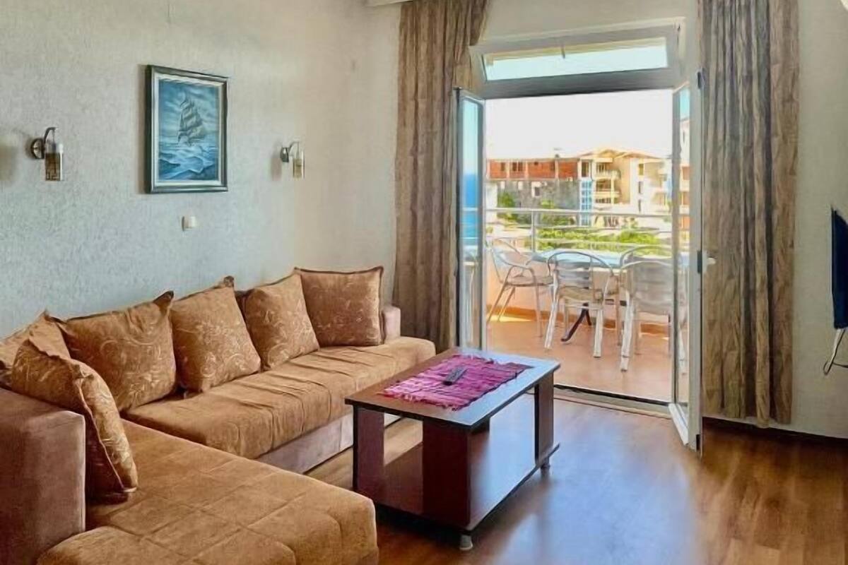 A living room with a brown sofa, a coffee table, and a painting on the wall. There is a sliding glass door leading to an outdoor area with a table and chairs.
