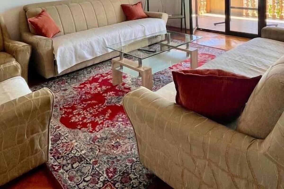 A living room with a sofa, armchairs, a glass coffee table, a painting, and a balcony with a view of buildings.