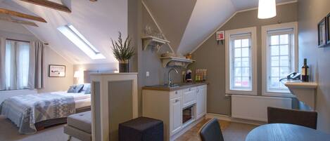 A compact, well-lit room with a bed, a small table, a chair, a kitchenette, and a window.